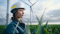 Female engineer in protective helmet at wind turbine farm, looking towards horizon through green crops. Sustainable energy and environmental concept.