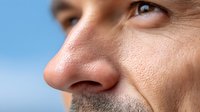 Close-up profile of middle-aged man's face showing nose, stubble, and eye against blue sky background, highlighting skin texture and facial features.