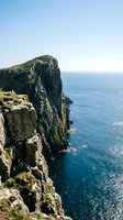 Dramatic rocky cliff edge with moss-covered stone formations overlooking calm blue ocean waters and distant coastline under clear sky. Perfect for travel and nature themes.