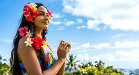 Young woman with long dark hair and medium brown skin tone wearing colorful flower lei garland and tropical floral crown, smiling outdoors against blue sky and palm trees, suitable for travel and summer campaigns.