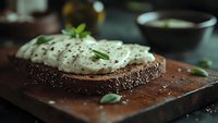 Fresh cream cheese spread on dark rye bread with herbs and black pepper on wooden cutting board for healthy breakfast or snack preparation.