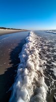 Ocean waves creating white foam along sandy beach shoreline under clear blue sky, dramatic perspective of surf meeting pristine coastline at sunrise.