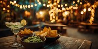 Margarita cocktail with guacamole and tortilla chips on wooden table with warm bokeh lights creating festive restaurant atmosphere for celebrations.