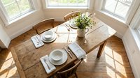Rustic wooden dining table set for two with white plates, napkins and fresh flower centerpiece in bright sunlit room with hardwood floors and large windows.