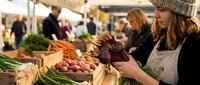 Young woman in knit hat shopping for fresh organic vegetables at bustling farmers market, selecting purple eggplant from colorful produce display.