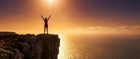 Man with arms raised standing on cliff edge at sunset overlooking ocean, celebrating success and achievement with golden sky and water backdrop.