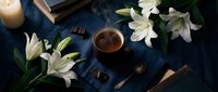 Cozy evening scene with white lilies, hot chocolate cup, books and candle on dark blue fabric creating peaceful atmosphere for relaxation and reading.