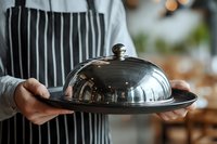 Waiter in striped apron holding silver cloche dome plate in upscale restaurant setting with blurred dining room background.