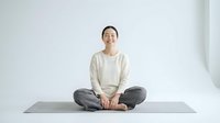 Young Asian woman in comfortable clothes sitting cross-legged on yoga mat in minimalist white studio space, peaceful meditation pose for wellness content.