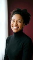 Happy young African American woman with natural curly hair smiling warmly in black turtleneck sweater against burgundy wall for lifestyle portraits.