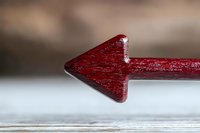Red wooden arrow pointing left on light background, macro shot with shallow depth of field, showing wood grain texture and directional guidance.