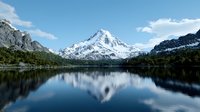 Majestic snow-capped mountain peak reflected in calm alpine lake waters, surrounded by rugged terrain and evergreen forest under bright blue sky with wispy clouds.