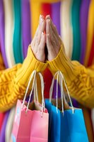 Female hands clasped in prayer position wearing yellow sweater with colorful shopping bags against rainbow striped background, expressing gratitude for purchases.