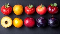 Fresh seasonal fruits arranged in rows on dark background - strawberries, kumquats, apples and plums showing variety of colors from yellow to deep purple.