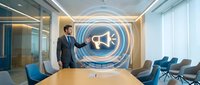 Professional businessman in dark suit presenting marketing strategy with megaphone hologram in modern conference room with blue chairs and glass walls.
