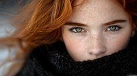 Close up portrait of young woman with red hair, freckles and green eyes wrapped in black scarf. Natural beauty concept with wind-blown hair and soft lighting.