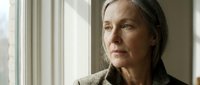 Senior woman with long gray hair and fair complexion gazes thoughtfully out window in natural daylight, wearing neutral cardigan for contemplative portrait use.