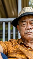 Senior Asian male farmer wearing straw hat and patterned shirt, sitting outdoors with serious expression. Agricultural worker portrait for farming, rural lifestyle, and agricultural industry concepts.