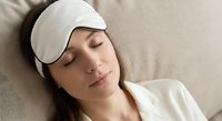 Young woman wearing white sleep mask resting peacefully on pillow in bed. Relaxation and healthy sleep concept for wellness and self-care routines.