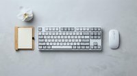 White mechanical keyboard and wireless mouse on gray concrete desk with bamboo notepad and orchid flower, minimalist workspace flat lay.