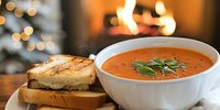 Homemade tomato soup with grilled cheese sandwich on wooden board, cozy fireplace and Christmas lights in background creating warm winter atmosphere.