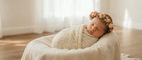 Peaceful newborn baby girl with floral headband sleeping wrapped in cream blanket on soft white fabric in warm natural light for portrait photography.