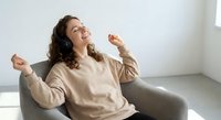 Young woman celebrating success with raised fists sitting on gray sofa in bright living room expressing joy and achievement in casual beige sweater.