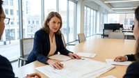 Professional businesswoman in navy suit leading meeting with colleagues in modern office conference room with city view windows.