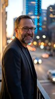 Confident middle-aged Caucasian businessman with glasses and beard smiling on urban balcony with blurred city lights background at dusk for corporate portraits.