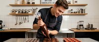 Male artisan chocolatier in black apron pouring melted chocolate onto mold in professional kitchen workshop. Craft confectionery production.