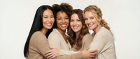 Four diverse women embracing in group hug wearing neutral sweaters against beige background. Friendship, sisterhood and female bonding concept.
