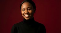 Young African American woman smiling warmly in black turtleneck against burgundy background for professional headshots and corporate marketing materials.