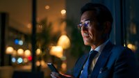 Professional Asian businessman in suit using smartphone in modern office at night with city lights background for corporate communication concepts.