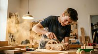 Woodworker sanding wooden toy car in workshop with dust particles floating in air, wearing glasses and dark shirt, surrounded by tools and materials for carpentry and craftsmanship projects.