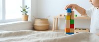 Child hands stacking colorful wooden blocks tower on bed in bright bedroom. Educational toy play activity for motor skills development and creativity.