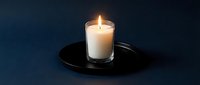 Lit white candle in clear glass holder on black plate against dark blue background, suitable for wellness, meditation, and home decor designs.