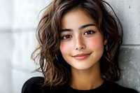 Young Asian woman with wavy brown bob hairstyle and natural makeup smiling confidently against concrete wall background wearing black sweater.