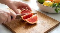 Person with medium skin tone slicing fresh grapefruit on wooden cutting board in bright kitchen setting. Food preparation and healthy eating concept.