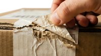 Caucasian male hand tearing open cardboard box with kraft paper tape, showing damaged packaging and shipping wear for logistics, delivery, and unboxing concepts.