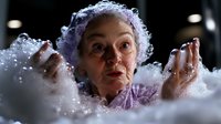 Senior woman with surprised expression emerging from white foam bubbles wearing purple clothing, dramatic lighting creates mysterious atmosphere for wellness and spa concepts.