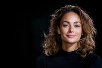 Young Mediterranean woman with wavy brown hair and natural makeup wearing black turtleneck smiling confidently against dark background. Professional headshot style portrait.