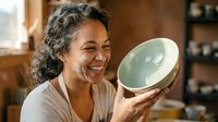 Middle-aged woman with curly dark hair and warm smile holding handmade ceramic bowl in pottery studio. Artisan craftswoman displaying glazed pottery work.