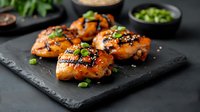 Glazed teriyaki chicken thighs garnished with sesame seeds and green onions served on black slate board. Asian fusion cuisine with sweet and savory marinade.