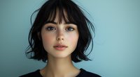 Young woman with short dark hair and bangs looking directly at camera against turquoise background, wearing dark top. Natural makeup and confident expression.