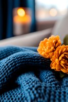 Cozy autumn composition with orange chrysanthemum flowers on blue knitted blanket, soft candlelight bokeh in background creates warm atmospheric mood.