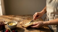 Woman in linen apron spreading butter on fresh homemade bread slice at rustic wooden table in warm kitchen light for breakfast preparation.
