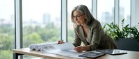 Mature businesswoman with gray hair and glasses reviewing documents at modern office desk near large windows with city view in bright workspace.