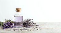 Glass bottle with cork stopper containing purple lavender essential oil surrounded by fresh and dried lavender flowers on rustic wooden surface for aromatherapy.