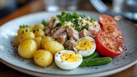Gourmet plate with flaked tuna, boiled potatoes, halved eggs, fresh tomato slices and green onions garnished with herbs and spices.