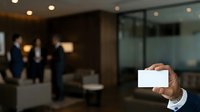 Hand holding blank white business card in modern office lobby with blurred colleagues in background, suitable for corporate branding, networking, and professional identity mockups.